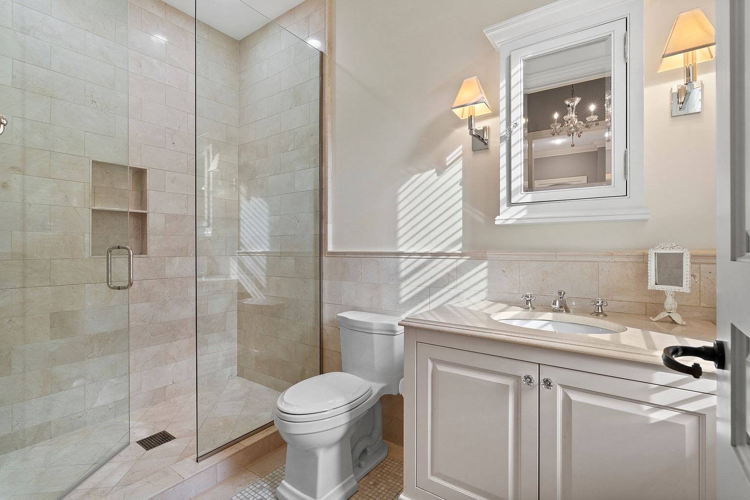 This bright and elegant bathroom features a walk-in shower enclosed by a large glass panel, showcasing light-toned stone wall tiles and a built-in recessed niche. A white vanity with a stone countertop and a matching medicine cabinet with integrated lighting creates a cohesive, polished look. The space is illuminated by soft wall sconces and natural light filtering through window blinds, contributing to a clean and sophisticated atmosphere.