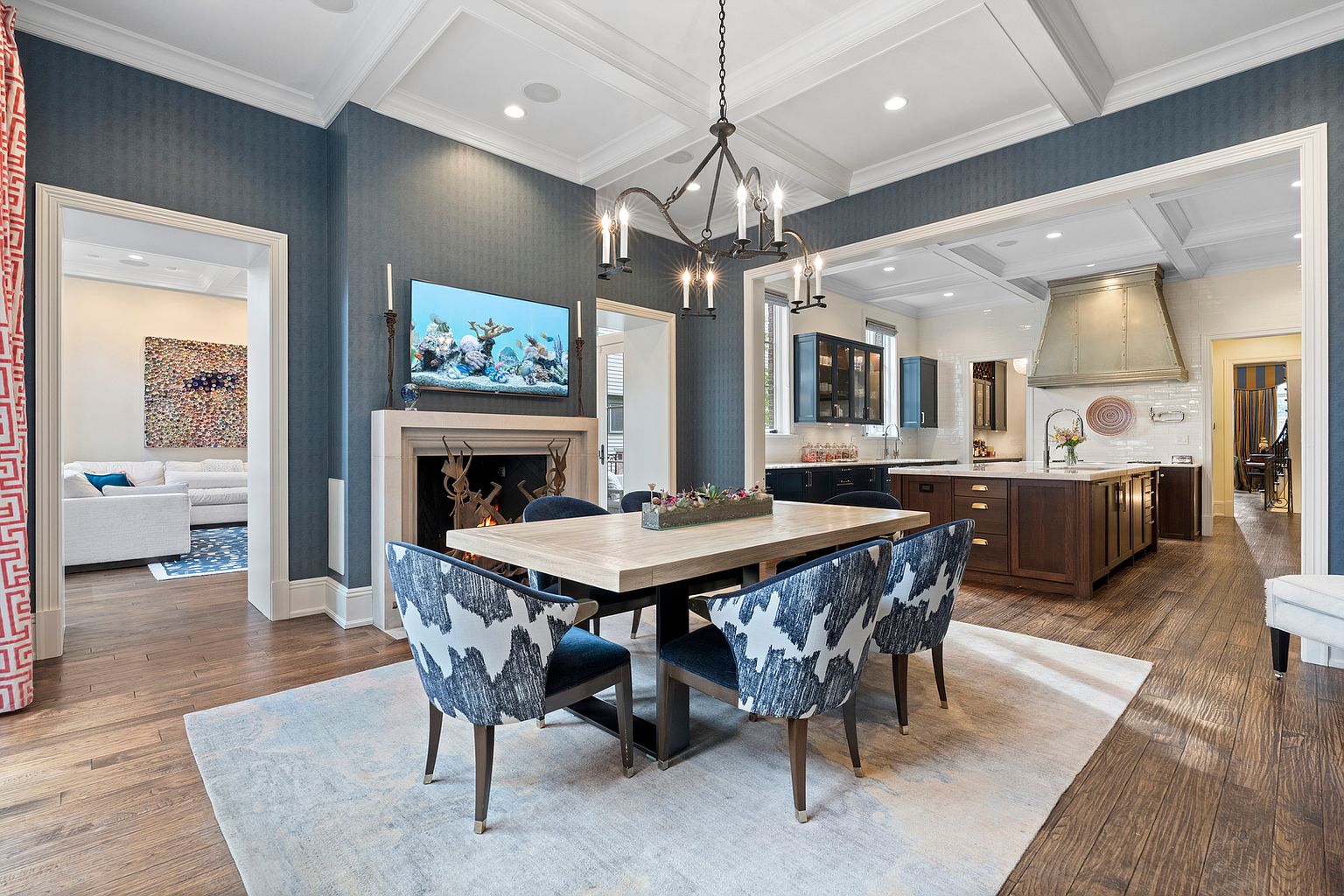 This elegant dining room features deep blue walls, a coffered ceiling, and a light wood dining table surrounded by patterned upholstered chairs. A stone fireplace with a decorative metal screen serves as a focal point, while an open-concept layout provides a clear view into the adjacent kitchen with its large island and metal range hood. The space is anchored by a light-colored area rug over dark wood flooring, creating a sophisticated and inviting atmosphere.