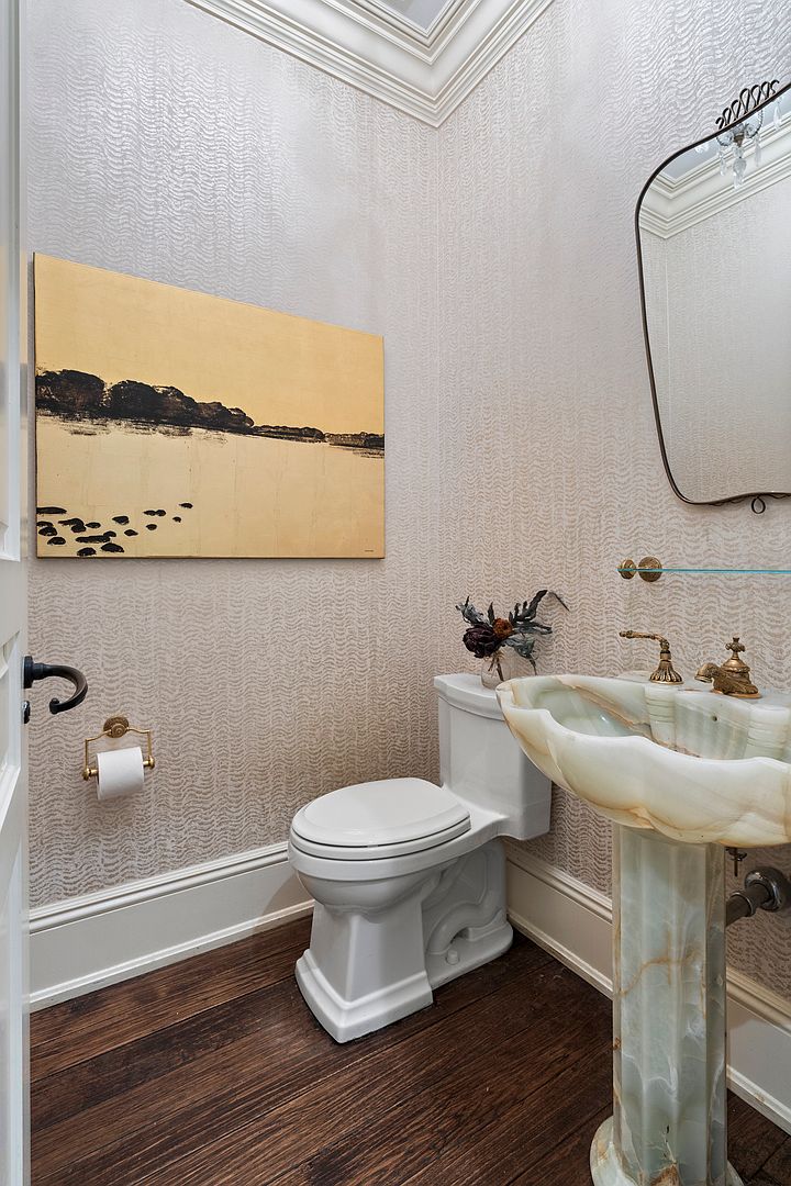 This elegant powder room features a unique pedestal sink crafted from translucent stone, paired with gold-toned fixtures and a decorative mirror. The walls are adorned with textured, neutral-toned wallpaper, complemented by dark wood flooring and a piece of abstract landscape art. The space is finished with crown molding and a classic white toilet, creating a sophisticated and compact atmosphere.