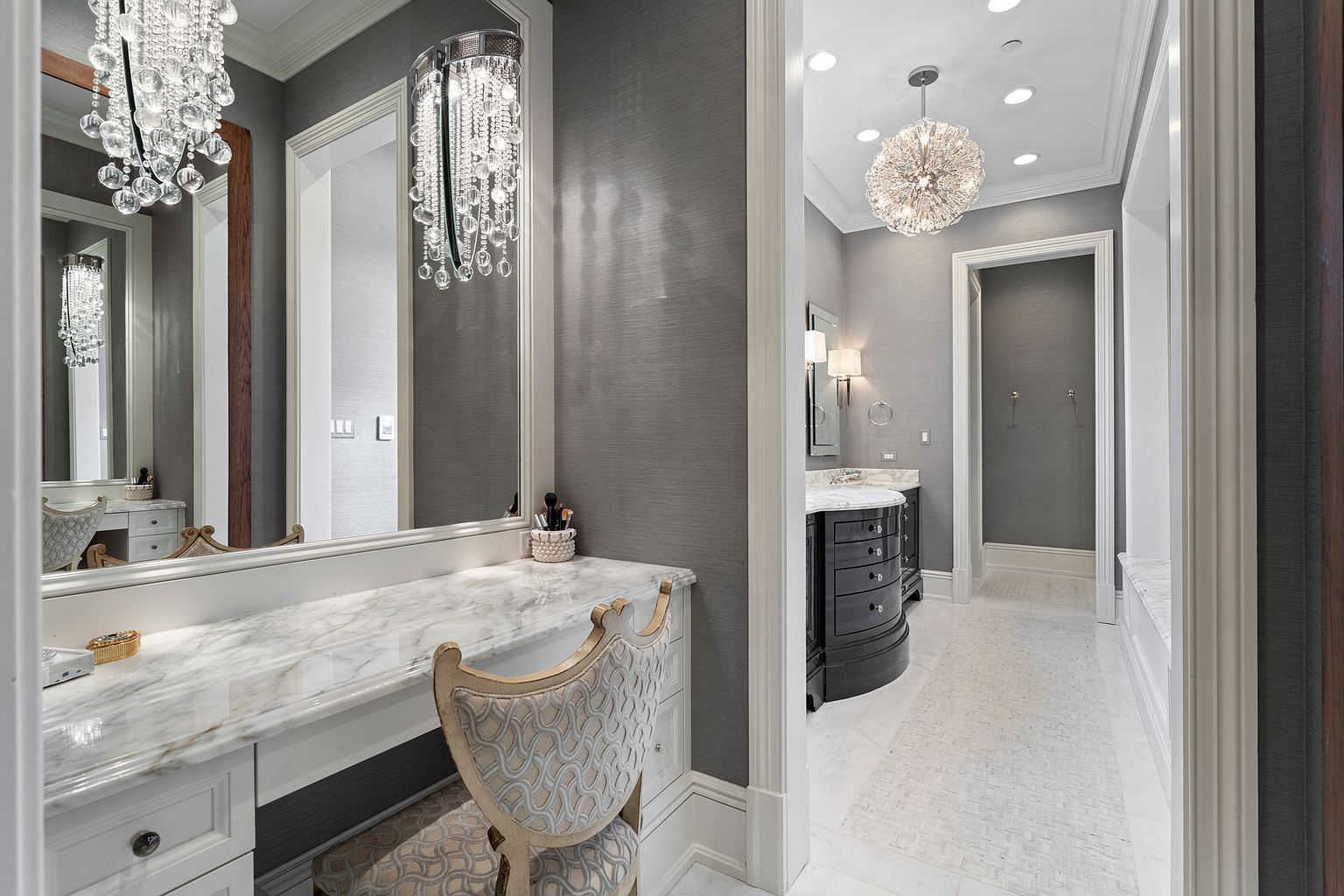 This elegant primary bathroom suite features a dedicated vanity area with a stone-topped desk, a patterned upholstered chair, and a large mirror accented by crystal chandelier lighting. The space transitions into a secondary area containing a dark-finished vanity and a hallway, all tied together by sophisticated gray textured wall coverings and light-toned stone flooring. The overall aesthetic is luxurious and refined, emphasizing high-end finishes and classic design elements.