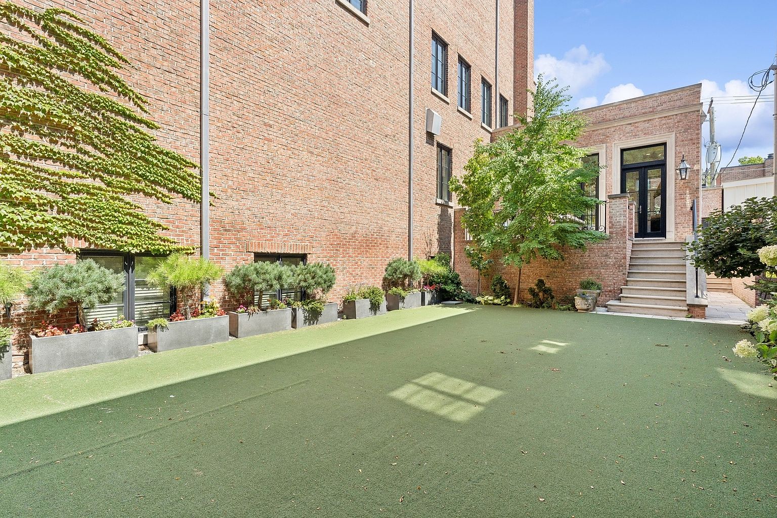 This private urban courtyard features a well-maintained synthetic turf lawn bordered by a series of modern, dark-toned planters filled with lush greenery. The space is framed by a tall, classic brick building with climbing ivy on one side and a raised stone patio area accessible by a set of stairs leading to a black-framed glass door. The scene offers a serene, secluded atmosphere, blending historic brick architecture with contemporary landscaping.
