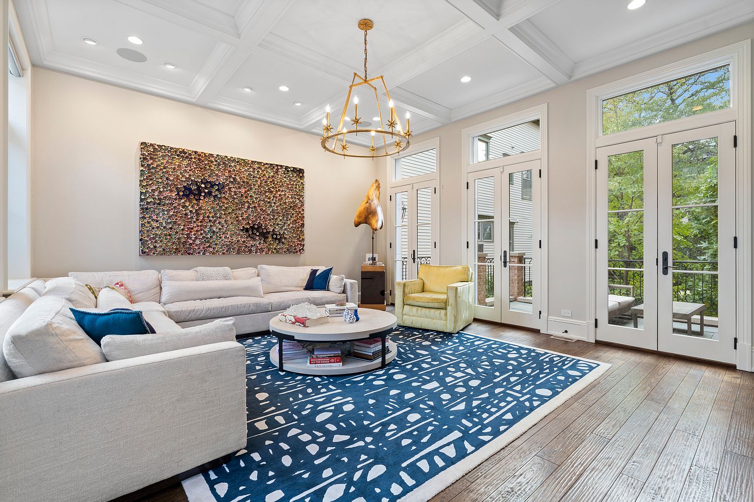 This bright and airy living room features a large, light-colored sectional sofa arranged around a circular coffee table on a bold, blue-and-white patterned area rug. A striking, multi-armed starburst chandelier hangs from a coffered ceiling, while tall glass doors provide abundant natural light and access to an outdoor balcony. The space is accented by a vibrant yellow armchair and a large, textured abstract art piece, creating a sophisticated and inviting atmosphere.