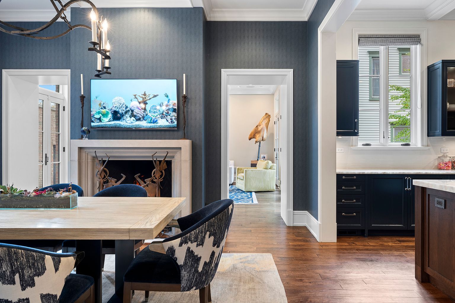 This elegant dining room features a light wood table surrounded by chairs with a distinctive black and white patterned fabric. A stone fireplace with a television mounted above serves as a focal point against a deep blue accent wall, while an open doorway leads to an adjacent living space. To the right, a glimpse of a kitchen with dark cabinetry and a window overlooking the exterior adds depth to the open-concept layout.