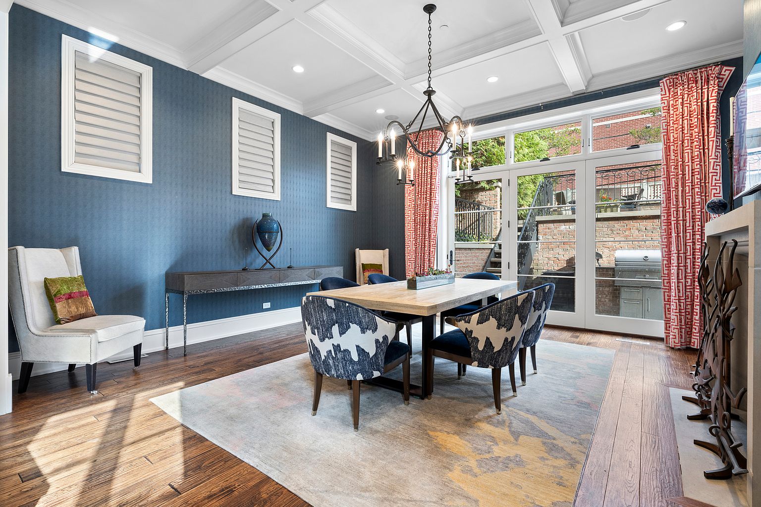 This elegant dining room features deep blue textured walls, a coffered ceiling, and rich wood flooring topped by a large, abstract-patterned area rug. A central wooden dining table is surrounded by chairs with bold, blue-and-white patterned upholstery, while a sleek console table sits against the wall. Large glass doors with red-patterned curtains provide a view of the outdoor patio, and a decorative chandelier hangs above the table, creating a sophisticated and inviting atmosphere.