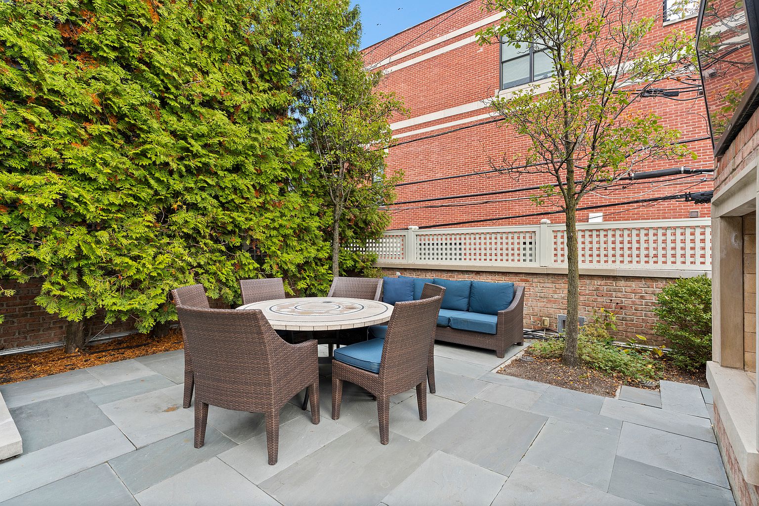 This private outdoor patio features stone pavers and a comfortable seating area with a round table and wicker-style chairs. A matching wicker sofa with blue cushions sits against a brick wall, complemented by mature trees and a white lattice fence. The space offers a cozy, secluded atmosphere perfect for outdoor dining or relaxation.