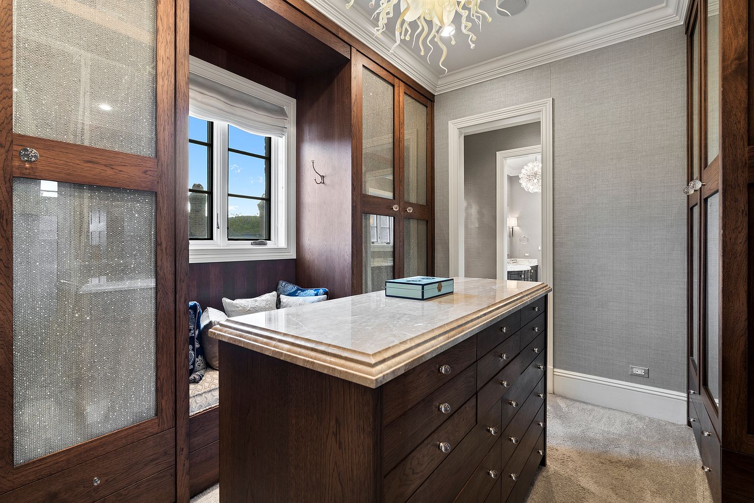 This luxurious walk-in closet features rich, dark wood cabinetry with glass-paneled doors and a central island topped with a smooth stone surface. A cozy window seat with patterned cushions is nestled between the storage units, while a decorative chandelier hangs from the ceiling. The room is finished with textured gray wall coverings and plush carpeting, creating an elegant and organized dressing space.