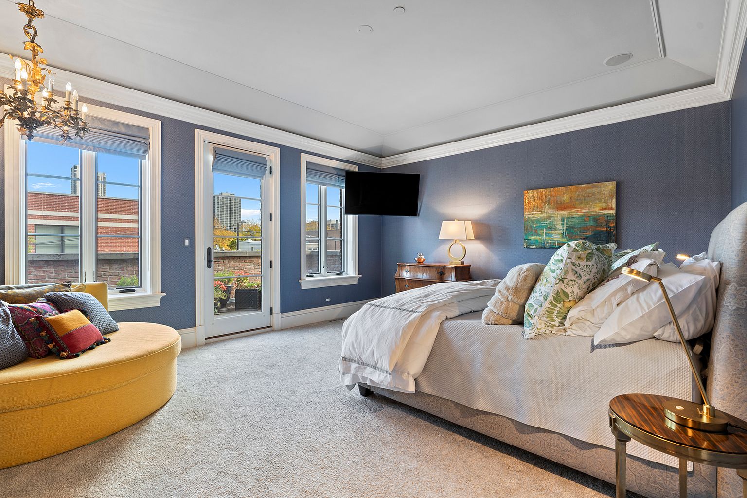 This elegant primary bedroom features deep blue textured walls, a plush light-colored carpet, and a sophisticated chandelier. The room is furnished with a comfortable bed adorned with patterned pillows, a circular yellow lounge chair, and a wooden dresser topped with a lamp. Large windows and a glass door provide ample natural light and access to an outdoor terrace, creating a bright and inviting atmosphere.