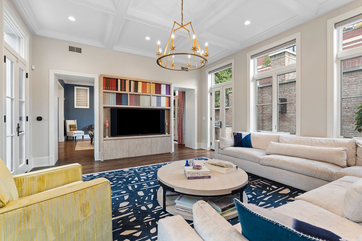 This bright and airy living room features a large, light-colored sectional sofa arranged around a circular coffee table on a bold, patterned blue and white rug. A custom light wood media unit with integrated shelving displays a colorful collection of books above a television, while a striking gold-toned chandelier serves as a focal point under the coffered ceiling. The room is bathed in natural light from several tall windows, creating an inviting and sophisticated atmosphere.