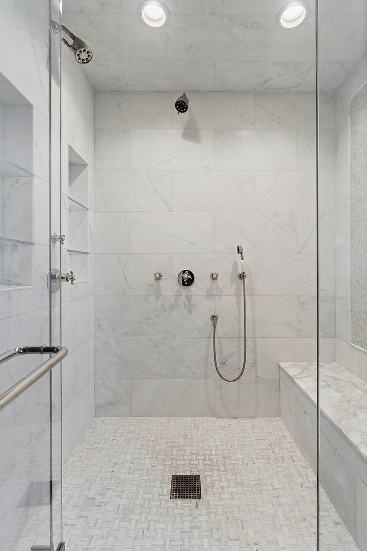 This spacious walk-in shower features light-toned stone wall tiles and a decorative mosaic tile floor. The enclosure includes a built-in stone bench, multiple shower heads, and integrated glass shelving, all framed by a clear glass door. The perspective is a direct, eye-level shot looking into the shower, highlighting the clean, luxurious aesthetic of the space.