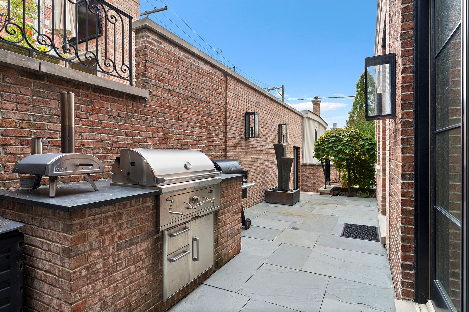 This outdoor patio space features a built-in brick kitchen island equipped with a stainless steel grill and a portable pizza oven. The area is paved with large stone tiles and enclosed by high brick walls, creating a private and sophisticated atmosphere. A modern water feature and wall-mounted lanterns add to the refined aesthetic of this urban outdoor living space.