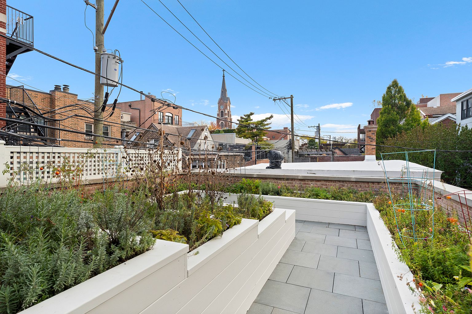 This rooftop terrace features a clean, modern design with raised white planter boxes filled with various herbs and plants. A central walkway paved with large, light-colored stone tiles leads the eye toward a scenic urban backdrop, highlighted by a prominent church steeple in the distance. The space is framed by a white lattice fence and offers an open, airy atmosphere perfect for outdoor relaxation.
