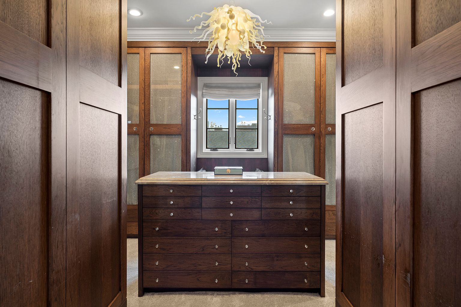This elegant walk-in closet features a central island with numerous drawers topped with a stone surface, framed by dark wood cabinetry with textured glass panels. A distinctive, sculptural chandelier hangs from the ceiling, casting a warm glow over the space, while a window centered between the cabinets provides natural light. The perspective is framed by two large wooden doors, creating a symmetrical and inviting view into the organized storage area.