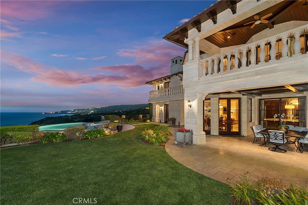 This luxurious Mediterranean-style estate features a sprawling rear patio and manicured lawn overlooking a dramatic coastal cliffside and ocean sunset. The home showcases elegant stone architecture, a covered outdoor living area with a dining set, and a vanishing-edge pool that blends seamlessly with the horizon. The perspective captures the grandeur of the property's outdoor amenities against a vibrant, twilight sky.