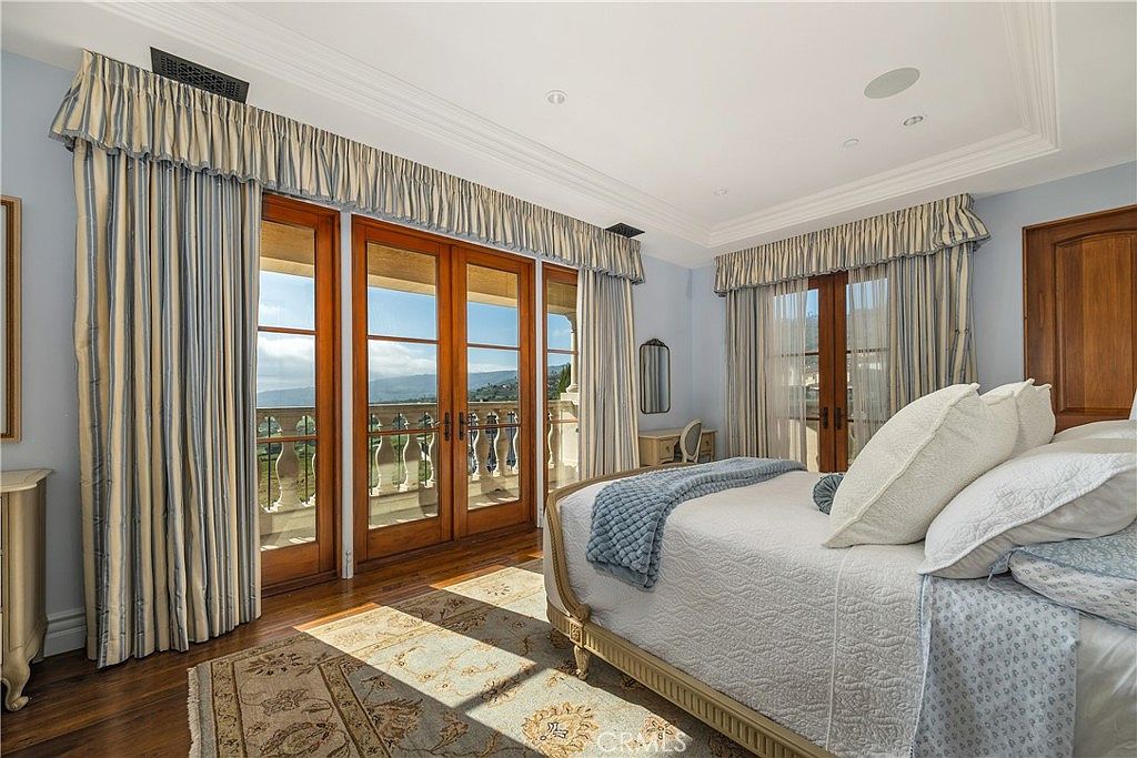 This elegant primary bedroom features a plush bed with layered bedding and a textured throw, positioned to face a set of tall glass doors that open onto a balcony. The room is decorated with soft blue walls, patterned curtains, and a large area rug, creating a serene and sophisticated atmosphere. The perspective captures the inviting sleeping area while highlighting the natural light streaming in from the outdoor view.