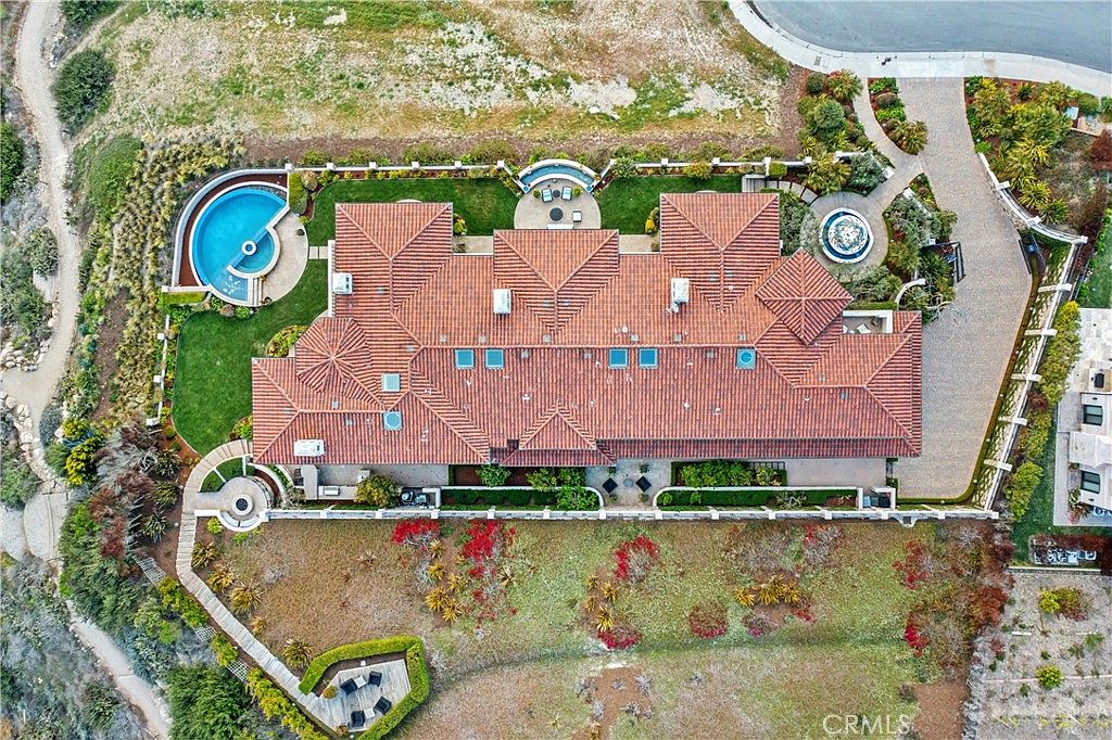 This high-angle aerial view captures a sprawling Mediterranean-style estate with a prominent red-tiled roof and multiple skylights. The property features a curved swimming pool, a circular spa, and a landscaped backyard with a stone-paved driveway leading to the front entrance. The surrounding grounds include manicured lawns, decorative shrubbery, and a natural hillside landscape, providing a sense of privacy and luxury.