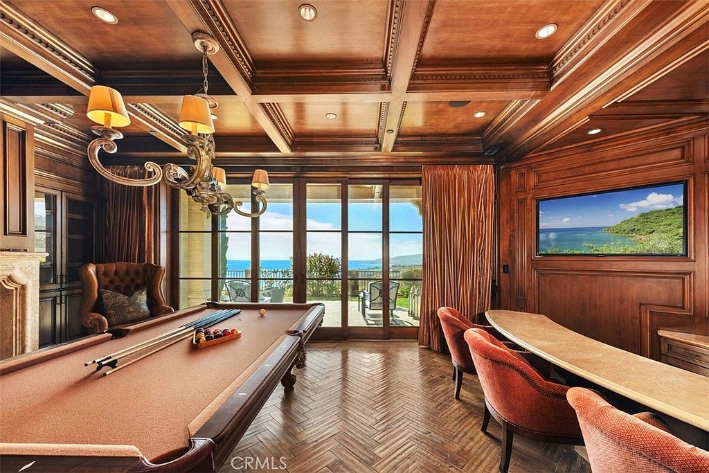 This sophisticated game room or study features rich wood-paneled walls, a coffered ceiling, and a classic herringbone-patterned wood floor. A billiard table sits in the center of the room, while a built-in bar counter with plush seating faces a wall-mounted television. Large glass doors provide a seamless transition to an outdoor patio with expansive ocean views, creating an elegant and relaxing atmosphere.