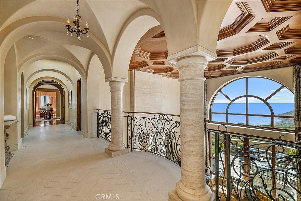 This elegant upper-level landing features a curved wrought-iron railing overlooking a lower floor, framed by grand stone columns and arched architectural details. The ceiling showcases intricate, geometric wood paneling, while a large arched window provides a sweeping view of the ocean horizon. The space is finished with light-toned stone flooring, creating a sophisticated and airy atmosphere.