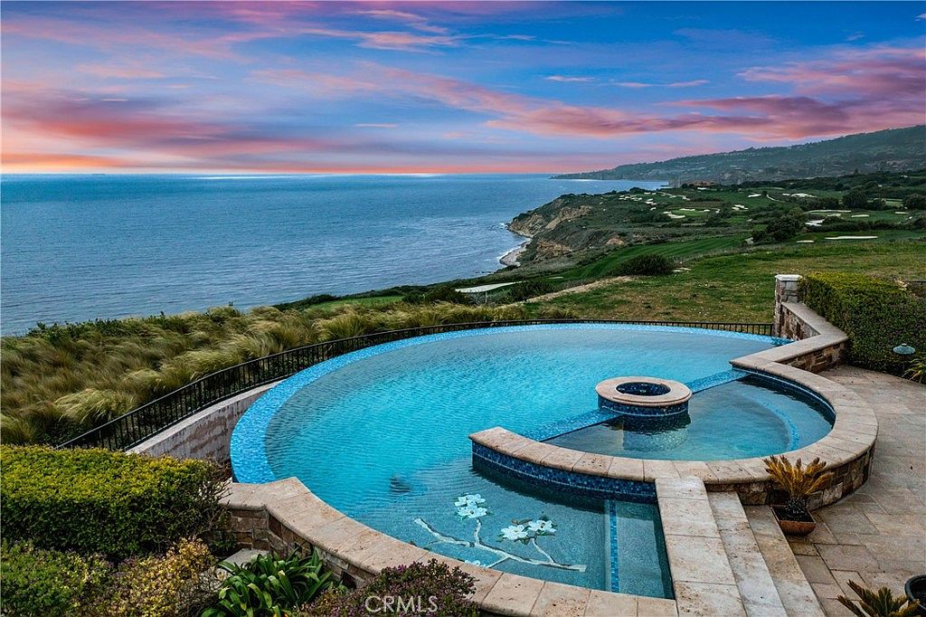 This stunning elevated perspective captures a luxurious infinity-edge pool and integrated spa overlooking a vast coastal landscape. The pool features intricate mosaic tile detailing at the bottom and is surrounded by stone pavers, set against a backdrop of the deep blue ocean and a vibrant sunset sky. The scene conveys a sense of high-end tranquility and exclusive coastal living.
