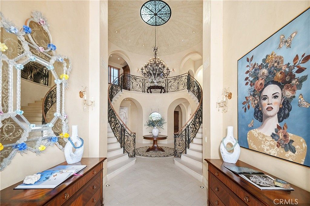 This grand foyer features a dramatic dual staircase with intricate wrought-iron railings that curve upward toward a central landing. The space is defined by high ceilings, a decorative domed ceiling with a circular skylight, and a large crystal chandelier as the focal point. Symmetrical wooden consoles flank the entrance, topped with decorative vases and artwork, creating a formal and luxurious atmosphere.