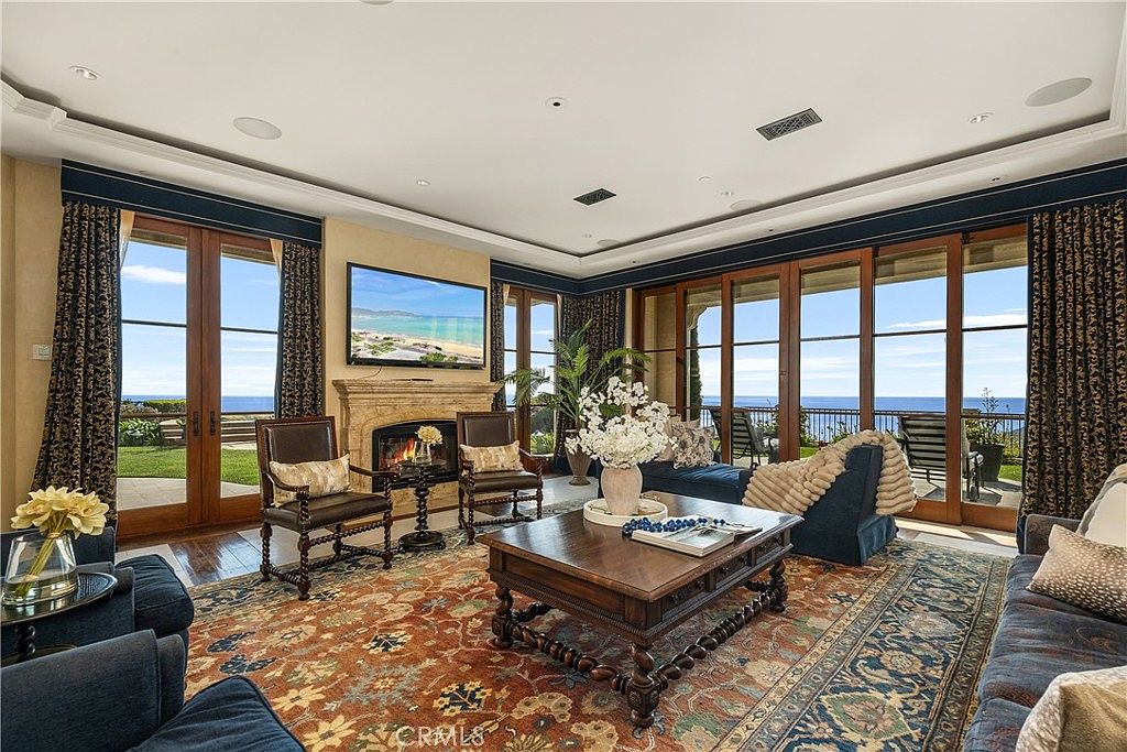 This elegant living room features a rich, patterned area rug, dark wood furniture, and a stone fireplace surround that serves as a central focal point. Large glass doors line the back wall, providing expansive views of the ocean and natural light throughout the space. The room is decorated with plush blue seating and sophisticated window treatments, creating a luxurious and comfortable atmosphere.
