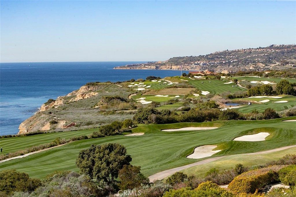 This expansive aerial view captures a prestigious coastal golf course set against the backdrop of the Pacific Ocean. The scene features meticulously manicured fairways, sand bunkers, and lush green landscaping that stretches toward the rugged coastline. The perspective provides a cinematic sense of scale, highlighting the prime location and serene atmosphere of this luxury property setting.