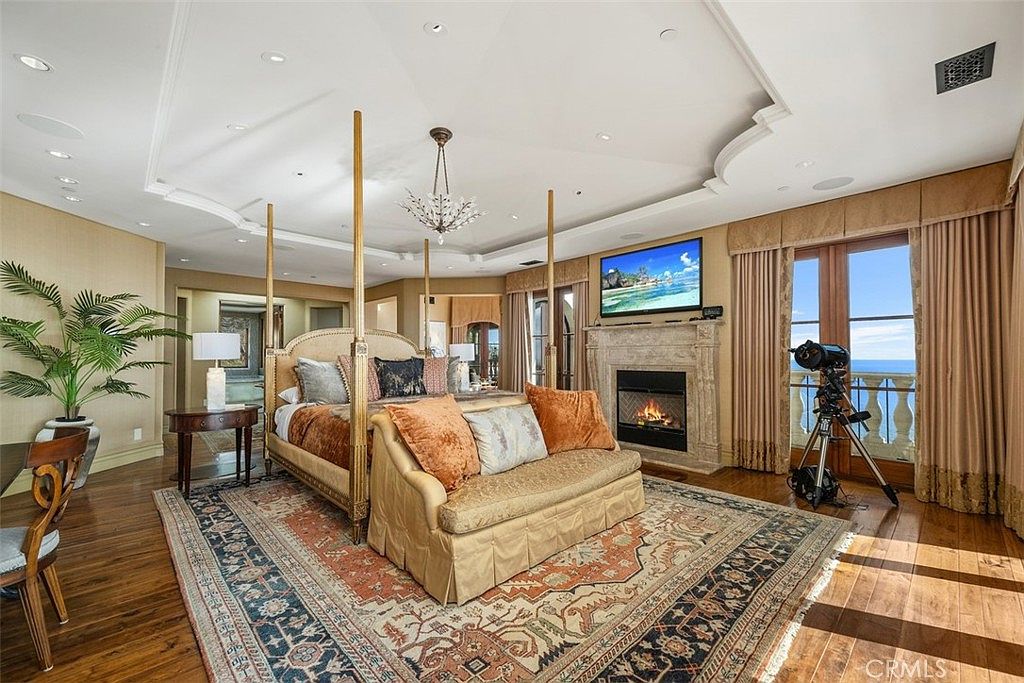 This luxurious primary bedroom features a grand four-poster bed as the centerpiece, set atop a large, intricately patterned area rug. A stone-clad fireplace with a mounted television sits between tall windows that offer a view of the ocean, while a plush bench sits at the foot of the bed. The room is accented by a crystal chandelier, recessed lighting, and warm-toned drapery, creating an elegant and sophisticated atmosphere.
