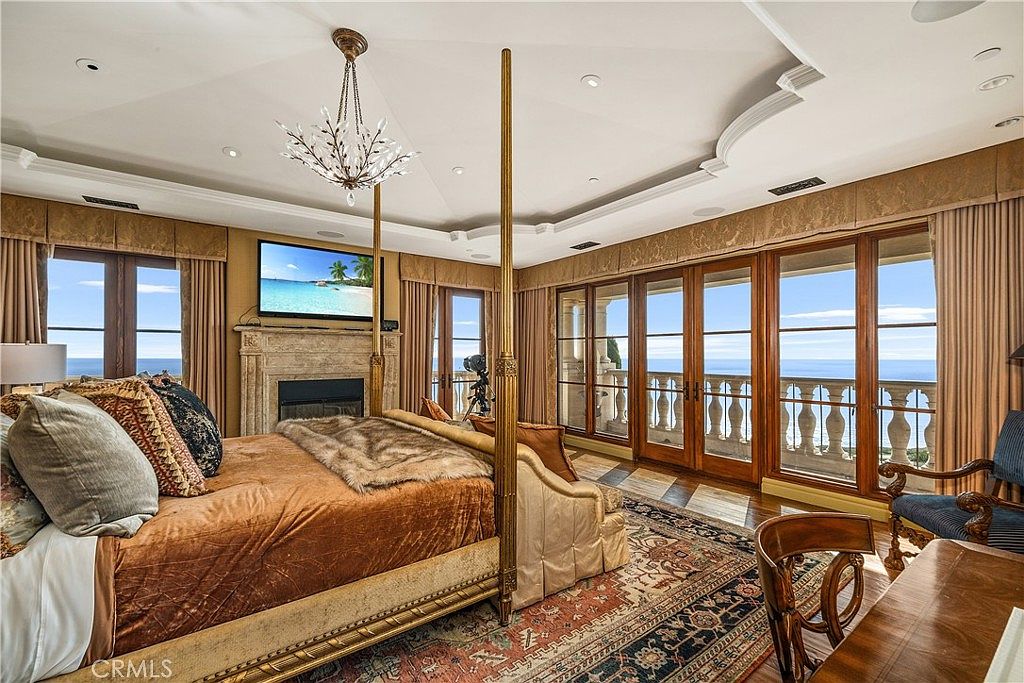 This luxurious primary bedroom features a prominent four-poster bed with a metallic frame, centered in the room beneath a tray ceiling with recessed lighting and a decorative chandelier. A stone-clad fireplace surround serves as a focal point, topped by a wall-mounted television, while expansive glass doors provide direct access to a balcony with ocean views. The space is finished with a patterned area rug, rich bedding, and a classic wooden desk, creating an elegant and serene atmosphere.