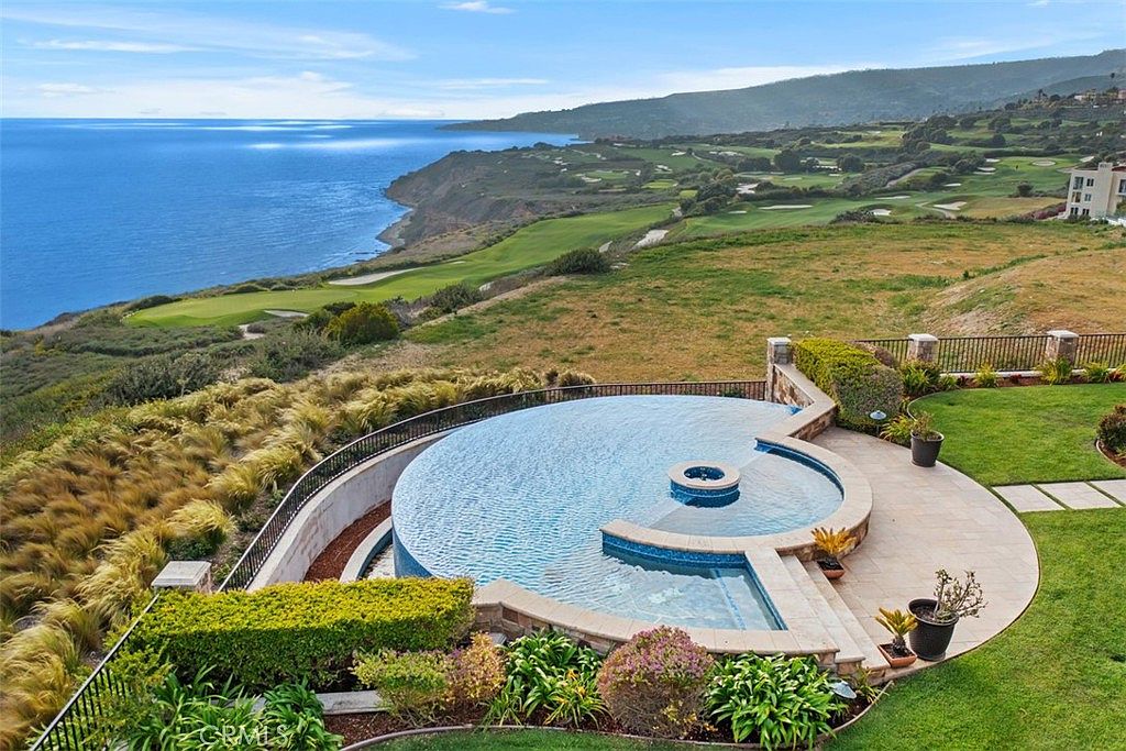 This high-angle aerial view captures a luxurious infinity-edge pool and integrated spa overlooking a vast coastal landscape. The pool area features a paved stone deck, lush landscaping, and a black metal fence, all set against a dramatic backdrop of rolling green hills and the deep blue ocean. The perspective emphasizes the property's prime location and seamless integration with the natural environment.