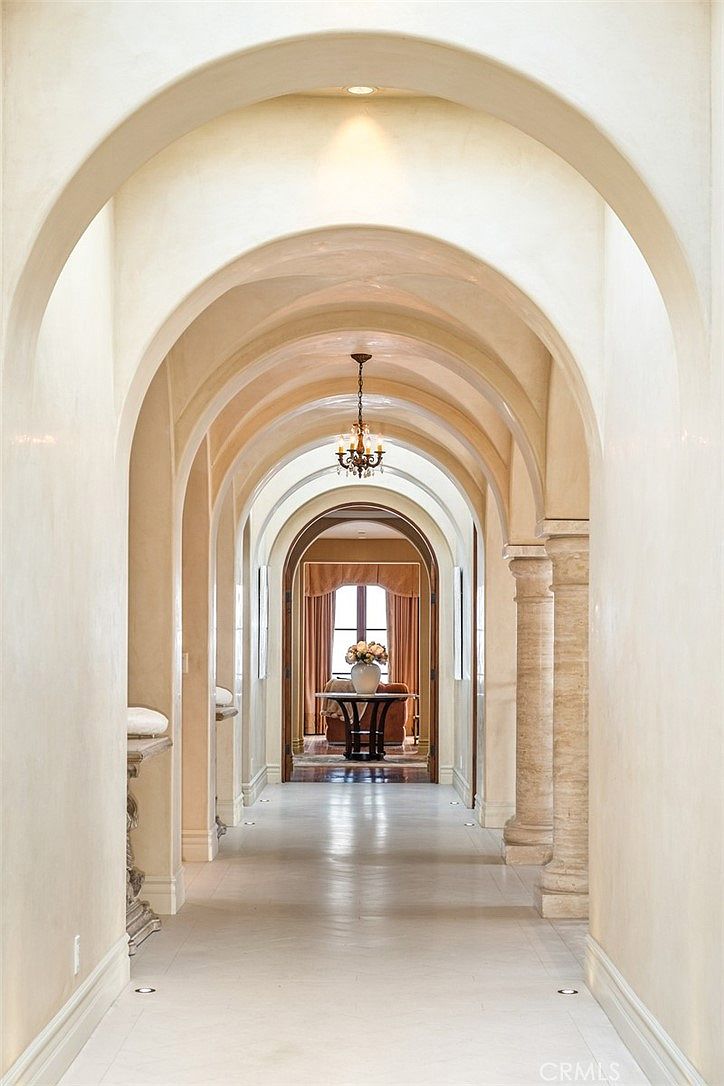 This elegant hallway features a series of dramatic, repeating arches that create a sense of depth and grandeur. The space is finished in a warm, neutral tone with polished light-colored flooring that reflects the soft overhead lighting. At the end of the corridor, a circular table with a floral arrangement sits in front of a window draped in soft curtains, providing a focal point for the perspective.