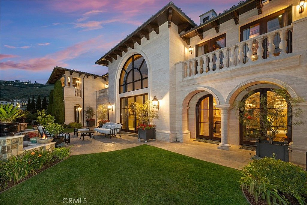 This luxurious Mediterranean-style estate features a sprawling stone patio overlooking a manicured lawn at dusk. The architecture is defined by arched doorways, a prominent two-story window feature, and a second-story balcony with stone balustrades. The warm glow of exterior lighting highlights the textured stone facade, creating an inviting and sophisticated atmosphere.