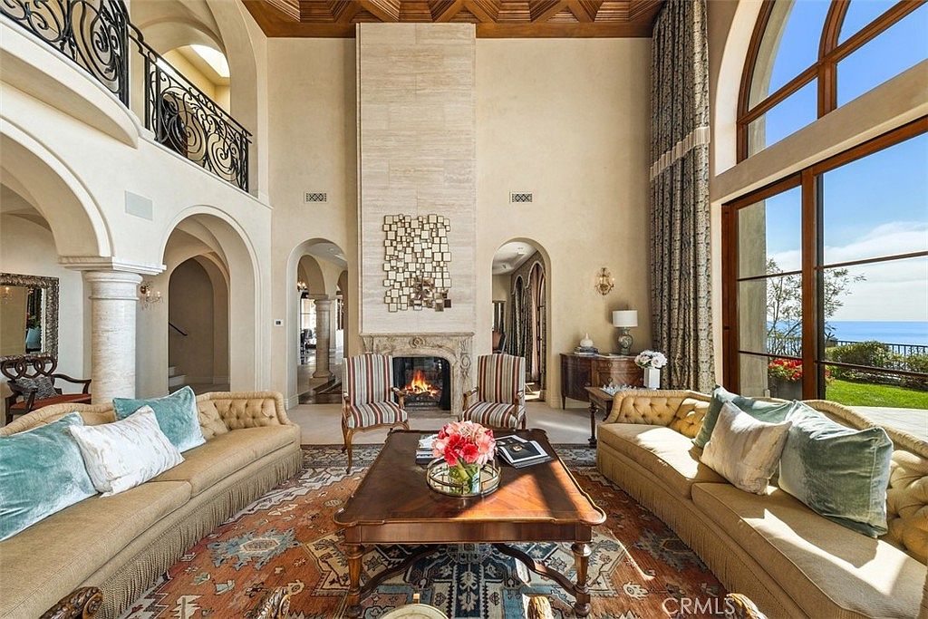 This grand living room features a soaring vaulted ceiling with exposed wood beams and a prominent stone-clad chimney breast centered between two arched doorways. The space is furnished with two large, plush sofas facing a central coffee table, complemented by a pair of striped accent chairs near the fireplace. Large windows provide an expansive view of the ocean, bathing the room in natural light and highlighting the elegant, traditional decor.
