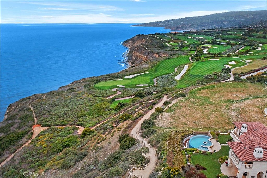 This high-angle aerial perspective captures a luxurious coastal estate perched on a cliffside overlooking the vast blue ocean. The property features a private swimming pool and spa, with a winding pathway leading through the natural landscape toward a sprawling golf course that hugs the rugged coastline. The scene emphasizes the dramatic topography and the exclusive, serene location of the residence.
