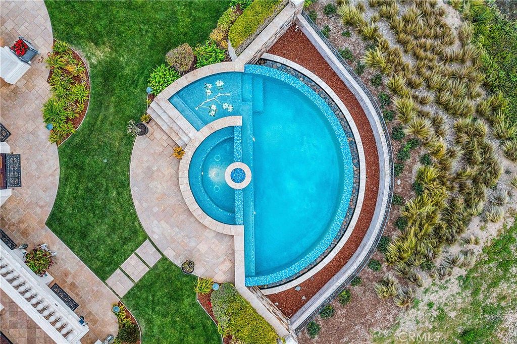This high-angle aerial view captures a custom-designed swimming pool and integrated spa set within a landscaped backyard. The pool features a unique semi-circular design with a tiled interior, surrounded by stone pavers, a manicured lawn, and a curved retaining wall. The surrounding area includes decorative landscaping and a naturalistic garden border, creating a serene and private outdoor retreat.