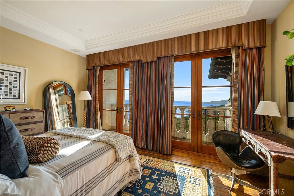 This elegant primary bedroom features warm-toned walls, rich wood flooring, and a patterned area rug that anchors the space. Large glass-paned doors open onto a private balcony, offering a stunning, unobstructed view of the ocean and coastline. The room is furnished with a comfortable bed, a classic desk setup, and a decorative arched mirror, creating a sophisticated and serene atmosphere.