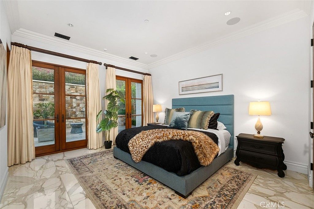 This primary bedroom features a plush bed with a blue upholstered headboard, accented by a leopard-print throw and dark nightstands with matching lamps. The room is anchored by a large, ornate area rug over light-toned stone flooring, while tall glass doors provide access to a private outdoor patio. The space is bright and airy, with white walls and recessed lighting creating a sophisticated and tranquil atmosphere.