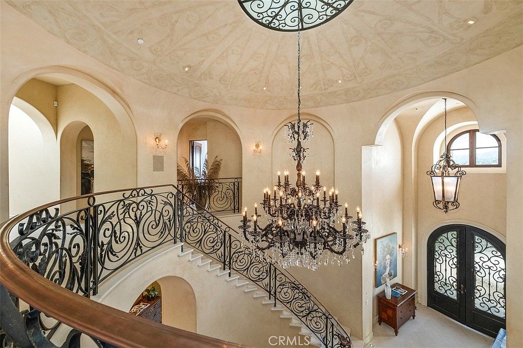 This grand foyer features a sweeping curved staircase with intricate wrought-iron railings that lead to an upper landing. A massive, multi-tiered crystal chandelier serves as the focal point, suspended from a domed ceiling adorned with decorative plaster molding. The space is characterized by arched doorways, neutral wall tones, and a sophisticated, Mediterranean-inspired architectural style.