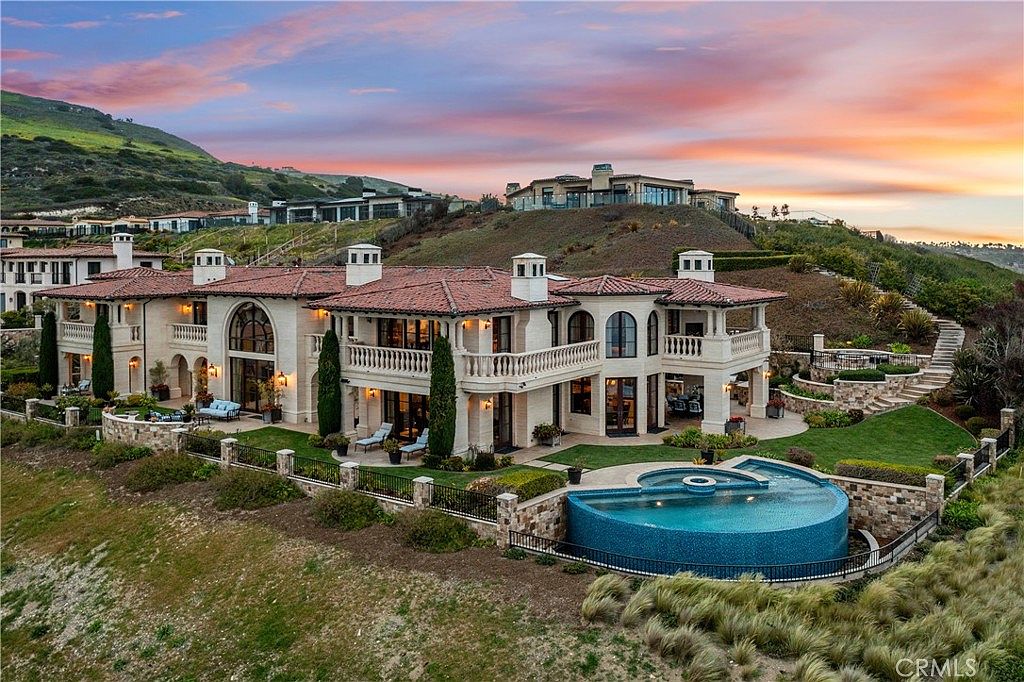 This expansive aerial view captures a luxurious Mediterranean-style estate perched on a hillside at sunset. The multi-level residence features a warm-toned stone facade, a classic tile roof, and multiple balconies overlooking a custom infinity-edge pool. The property is surrounded by landscaped grounds and rolling hills, creating a serene and high-end atmosphere.