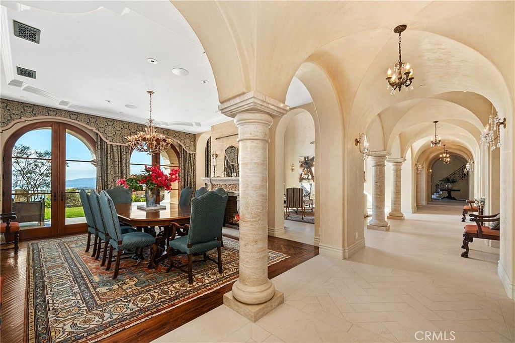 This elegant dining room features a large wooden table surrounded by upholstered chairs, set atop a detailed patterned rug on dark wood flooring. The space is defined by grand arched openings and stone columns that lead into a long, vaulted hallway, creating a sense of architectural depth and luxury. A crystal chandelier hangs above the table, while large arched glass doors provide a view of the outdoor landscape, bathing the room in natural light.