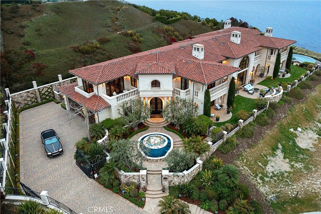 This high-angle aerial view captures a sprawling Mediterranean-style estate perched on a coastal cliffside. The residence features a complex terracotta tile roof, light-toned stone walls, and a grand circular driveway with a decorative fountain centerpiece. Lush landscaping, a private pool area, and expansive ocean views define this luxurious property, which exudes an air of exclusivity and architectural elegance.