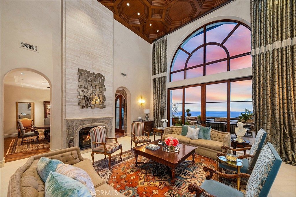 This grand living room features a dramatic coffered wood ceiling and a tall stone-clad chimney breast centered on the wall. Large arched windows provide an expansive view of the ocean at sunset, while a vibrant patterned rug anchors the seating area composed of plush sofas and armchairs. The space exudes a luxurious, formal atmosphere with elegant drapery and a sophisticated architectural design.