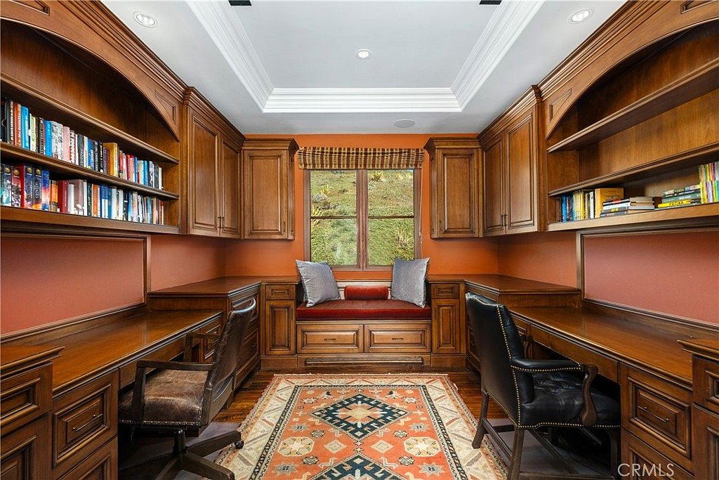 This sophisticated home office features extensive built-in wooden cabinetry and shelving flanking a central window with a built-in bench seat. Two matching desks are positioned on opposite sides of the room, creating a symmetrical workspace layout centered around a patterned area rug. The warm wood tones and orange wall accents provide a classic, scholarly atmosphere suitable for a professional study.