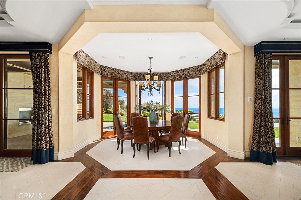 This elegant dining room features a central round table surrounded by six high-backed leather chairs, positioned under a decorative chandelier. The room is defined by a hexagonal floor pattern with dark wood borders and light-colored tile, framed by large windows that offer expansive ocean views. Ornate patterned valances top the windows, and the space is flanked by heavy, dark curtains, creating a formal and sophisticated atmosphere.