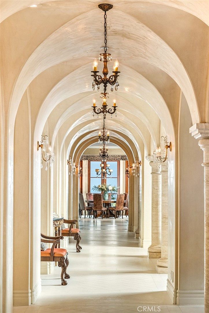 This grand, arched hallway features a series of repeating barrel-vaulted ceilings and classical columns, creating a dramatic sense of depth and perspective. Ornate multi-tiered chandeliers hang from the center of each arch, casting a warm glow over the light-toned stone flooring and elegant wooden chairs placed along the walls. The hallway leads the eye toward a bright dining area at the far end, emphasizing the luxurious and architectural scale of the home.