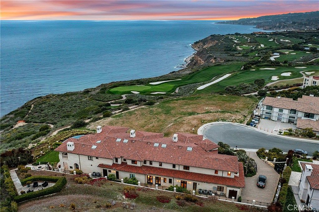 This high-angle aerial shot captures a sprawling Mediterranean-style estate perched on a coastal cliffside overlooking the ocean and a golf course. The home features a multi-gabled roof with terracotta tiles, a paved motor court, and expansive views of the rugged coastline. The perspective emphasizes the property's prime location, showcasing the seamless integration between the luxury residence and the surrounding natural landscape.