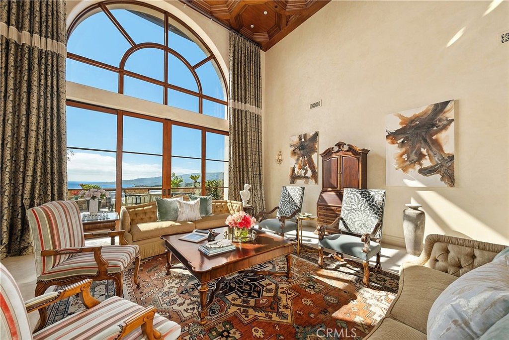 This elegant living room features a dramatic, multi-paneled arched window that frames a scenic ocean view. The space is anchored by a large, intricately patterned area rug and furnished with a mix of classic upholstered seating, including a tufted sofa and patterned armchairs. A coffered wood ceiling adds architectural depth, while two large abstract paintings and a traditional wooden cabinet provide sophisticated decorative accents.