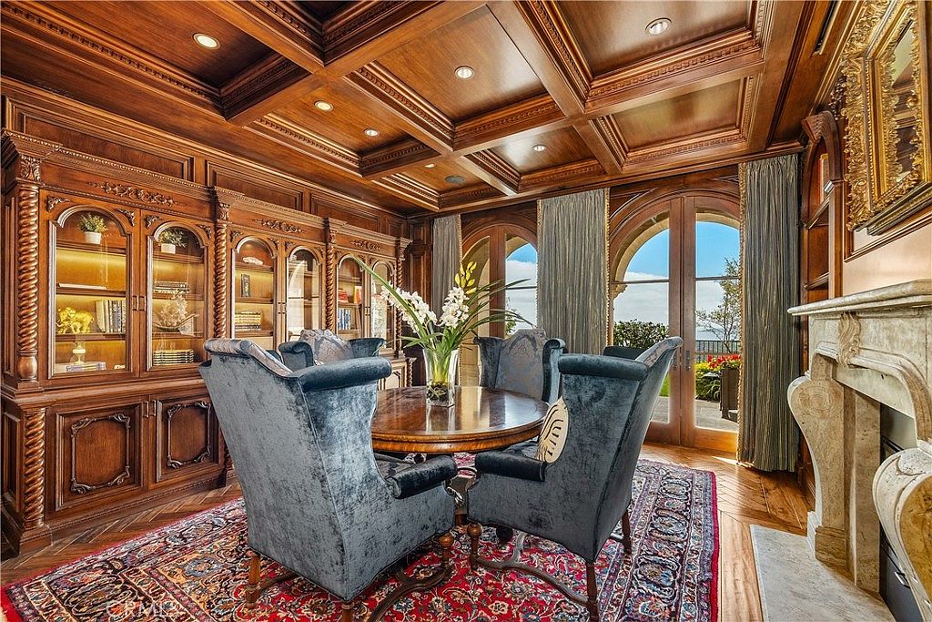 This sophisticated study features rich wood-paneled walls with built-in cabinetry and a coffered ceiling, creating an atmosphere of classic elegance. A round table sits in the center surrounded by plush, velvet-upholstered chairs, while a patterned area rug adds warmth to the space. Large glass doors provide natural light and a view of the outdoors, complemented by a stone fireplace surround on the right.
