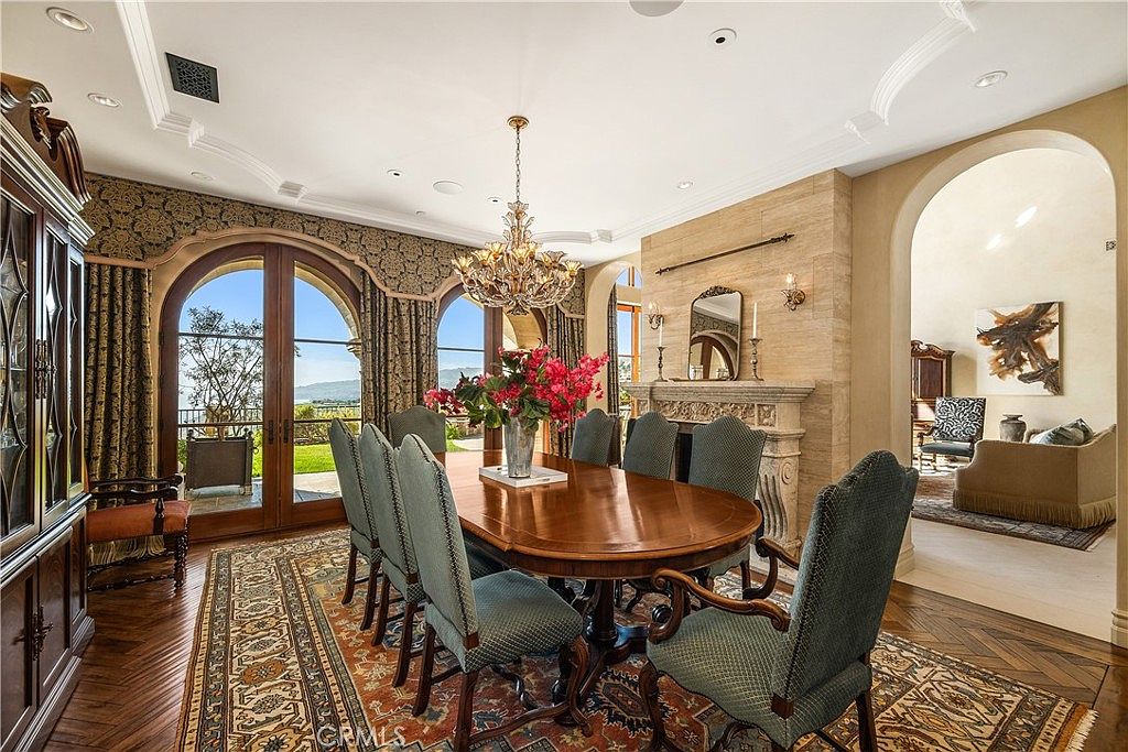 This elegant dining room features a large wooden table surrounded by upholstered chairs, set atop a detailed patterned area rug on dark wood flooring. A prominent stone-clad chimney breast serves as a focal point, complemented by a crystal chandelier and arched glass doors that provide a view of the outdoor landscape. The space is framed by an arched entryway leading to an adjacent room, creating a sophisticated and open atmosphere.