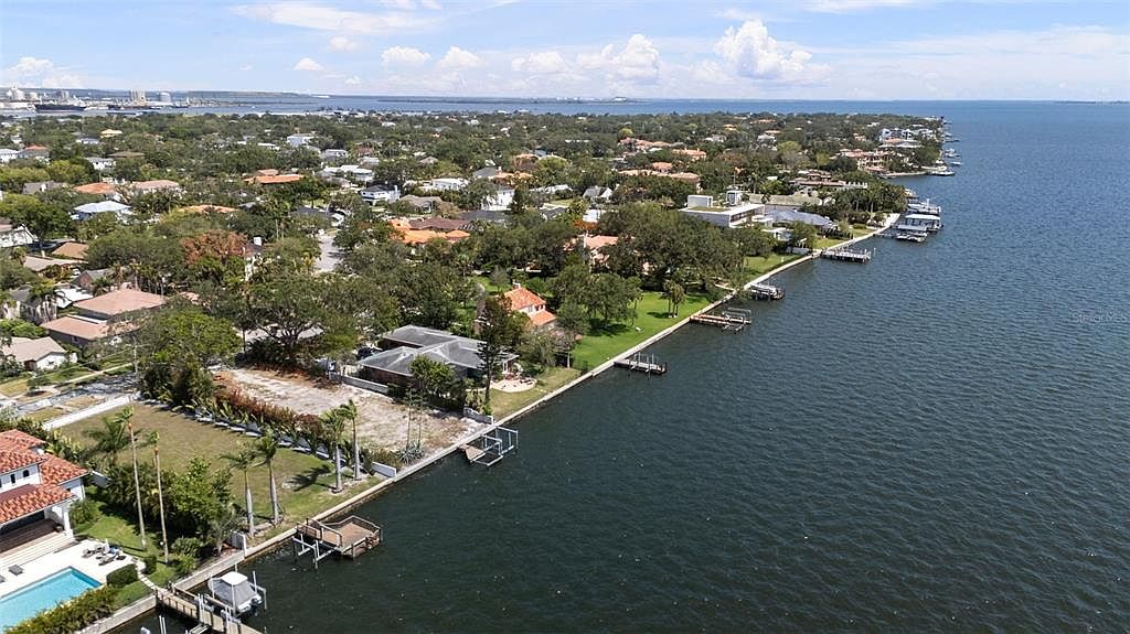 This aerial shot showcases a waterfront property with a clear view of the bay. The neighborhood features lush greenery and well-maintained homes, some with private docks. The overall impression is one of luxury and serene coastal living.