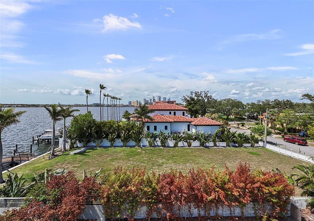 This aerial view showcases a luxurious waterfront property with a red tile roof and white exterior walls. Lush landscaping surrounds the house, and a private dock extends into the water. The image captures the serene setting and highlights the property's desirable location and upscale features.