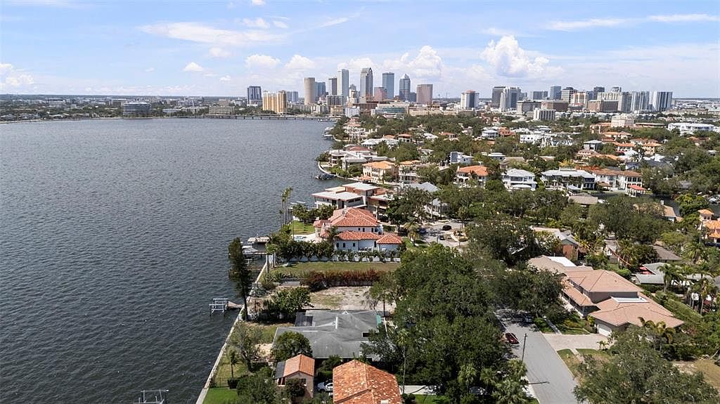 This aerial view showcases a waterfront property with a stunning cityscape backdrop. The image captures a mix of residential homes with varying architectural styles, lush greenery, and a serene body of water. The overall impression is one of luxury and prime real estate location.